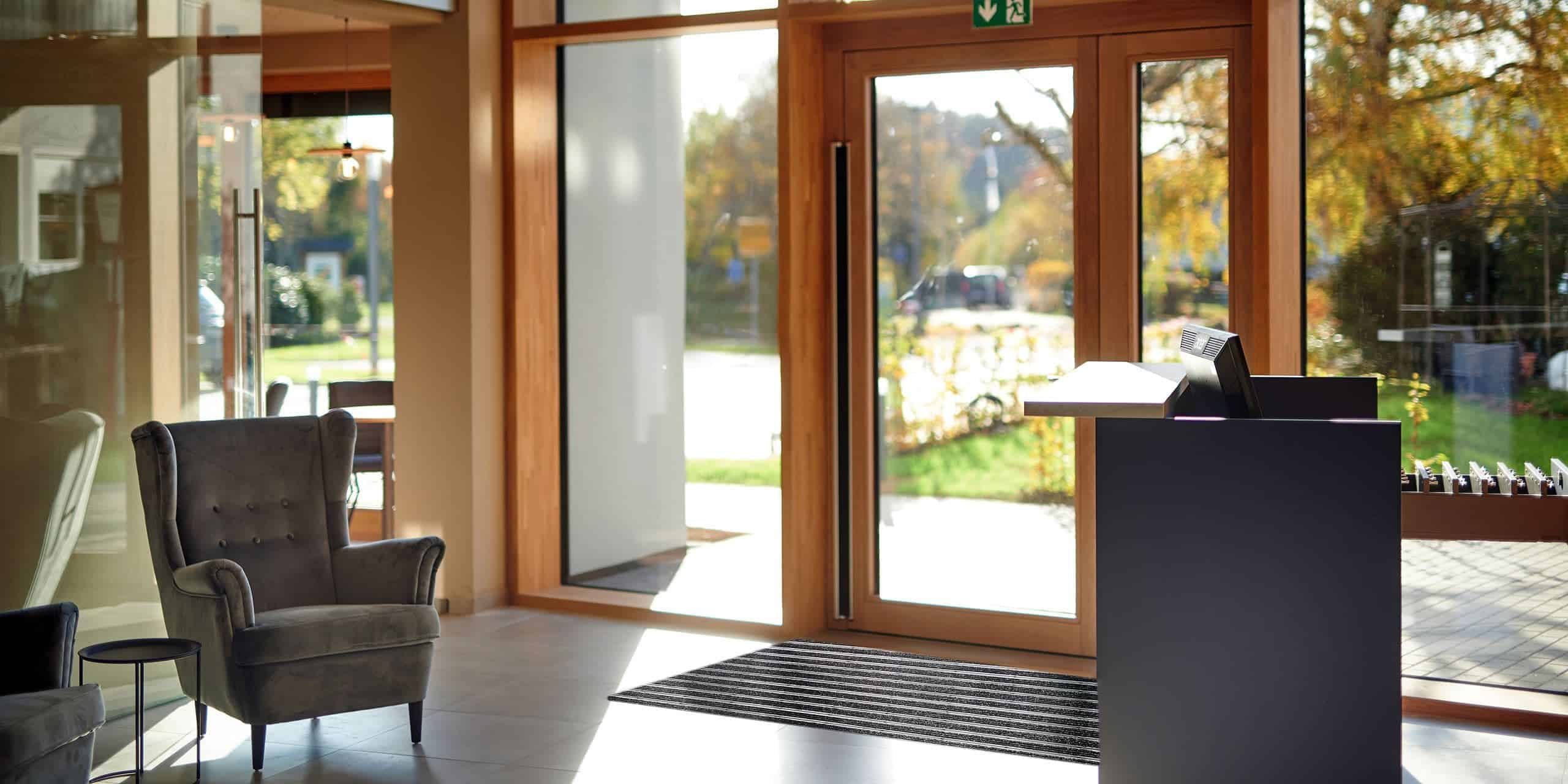Blick aus der Lobby von Kucher's Genusshotel in der Eifel in den grünen Garten, blauer Himmel und warmes Licht. Einladende Verbindung von Innen und Außen.